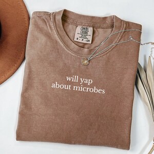 May include: A brown Comfort Colors t-shirt with the text "will yap about microbes" in white. A silver necklace with a small pendant is draped across the shirt. The shirt is folded and lies on a white surface.