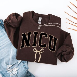 Op de afbeelding: Een bruine sweatshirt met het woord "NICU" in zwarte blokletters met een luipaardprint. Onder de tekst staat een crèmekleurige strik. Een zilveren ketting is over de sweatshirt gedrapeerd. De sweatshirt is op een spijkerbroek gevouwen.