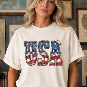 Könnte beinhalten: Ein cremefarbenes Kurzarm-T-Shirt mit dem Schriftzug USA in einer großen, fetten Schriftart. Die Buchstaben haben eine abgenutzte Jeans-Textur mit rot-weißem Streifenmuster und weißen Sternen, die das Design der amerikanischen Flagge nachahmen.