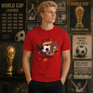 May include: A red short-sleeved t-shirt featuring a graphic print of a football, the German flag, a black eagle, the Brandenburg Gate, and a gold trophy. The text GERMANY 20 is printed below the central graphic in a bold, dark font.