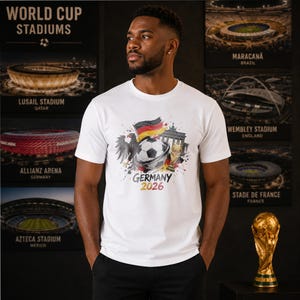 May include: A white short-sleeved t-shirt featuring a graphic print of a soccer ball, the German flag, an eagle, the Brandenburg Gate, and a gold trophy. The text "GERMANY 2026" is printed below the graphic in black and yellow lettering.