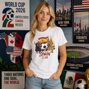 May include: A white t-shirt featuring a graphic print of a bull, a soccer ball, the Spanish flag, the Sagrada Familia, and a gold trophy. Below the graphic, the text reads "SPAIN 2026" in red and gold lettering, centered on the chest area.