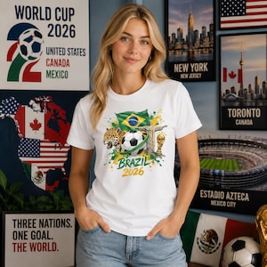 May include: A white short-sleeve t-shirt featuring a graphic print of a soccer ball, a jaguar, the Brazilian flag, the Christ the Redeemer statue, and a gold trophy with the text Brazil 2026 centered on the chest area of the garment.