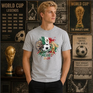 Pode incluir: Uma t-shirt cinzenta mesclada de manga curta com uma impress&atilde;o gr&aacute;fica de uma bola de futebol, a bandeira do M&eacute;xico, uma &aacute;guia-real e uma pir&acirc;mide. O design inclui o texto MEXICO 2026 em letras verdes e vermelhas na zona do peito.