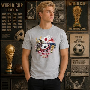Puede incluir: Una camiseta gris jaspeado de manga corta con un estampado gr&aacute;fico de un casco de samur&aacute;i, un bal&oacute;n de f&uacute;tbol, un trofeo dorado, la bandera de Jap&oacute;n y el monte Fuji, con el texto "JAPAN 2026" impreso en rojo bajo la ilustraci&oacute;n.