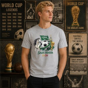 Pode incluir: Uma t-shirt cinzenta mesclada com uma impress&atilde;o gr&aacute;fica de uma bola de futebol, um cavalo branco, a bandeira da Ar&aacute;bia Saudita, um trof&eacute;u dourado e uma torre de rel&oacute;gio. O texto "SAUDI ARABIA 2026" est&aacute; impresso em verde e vermelho.