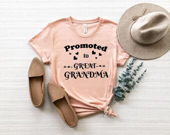 May include: A light pink t-shirt with the text "Promoted to Great Grandma" in black. The shirt is styled with a pair of brown flats, a beige hat, and greenery.