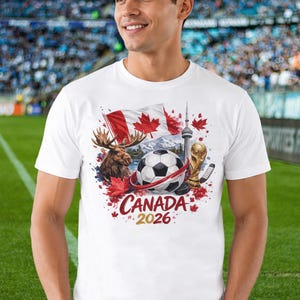 May include: A white short-sleeved t-shirt featuring a graphic print of a soccer ball, a moose, the Canadian flag, the CN Tower, and a gold trophy. The design includes red maple leaves and the text "CANADA 2026" printed in red and gold lettering.