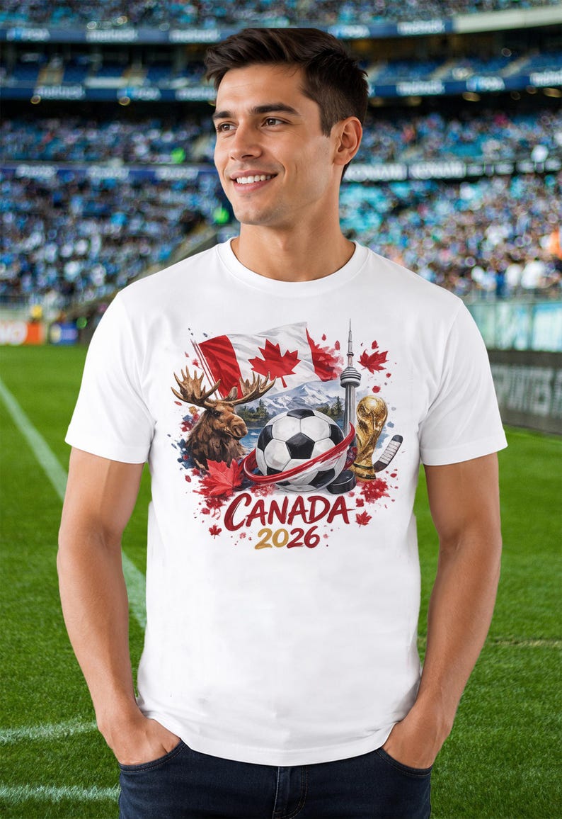 May include: A white short-sleeved t-shirt featuring a graphic print of a soccer ball, a moose, the Canadian flag, the CN Tower, and a gold trophy. The design includes red maple leaves and the text "CANADA 2026" printed in red and gold lettering.