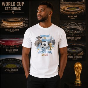 May include: A white short-sleeved t-shirt featuring a graphic print of a soccer ball, the Argentina flag, a cowboy figure, the Obelisco de Buenos Aires, and a gold trophy. The text reads Argentina 2026 in blue and gold lettering on the front.