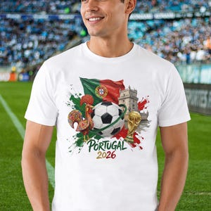 May include: A white short-sleeved t-shirt featuring a graphic print of the Portuguese flag, a soccer ball, a rooster, the Bel&eacute;m Tower, and a gold trophy. The text "PORTUGAL 2026" is printed in green and gold lettering below the central graphic design.