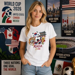 May include: A white t-shirt featuring a graphic print of a soccer ball, a tiger, the South Korean flag, a traditional building, and a gold trophy. Below the graphic, the text reads KOREA 2026 in red and gold lettering on the front of the shirt.