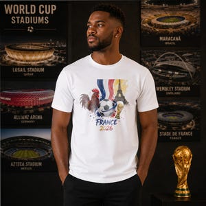 Puede incluir: Una camiseta blanca de manga corta con un estampado gr&aacute;fico de un gallo, un bal&oacute;n de f&uacute;tbol, la Torre Eiffel y un trofeo dorado, con el texto France 2026 mostrado debajo del dise&ntilde;o en letras azules y doradas.