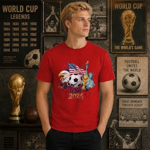 May include: A red short-sleeved t-shirt featuring a graphic print of a bald eagle, a soccer ball, the American flag, the Statue of Liberty, and a gold trophy. The text USA 2026 is printed in gold below the central graphic elements on the chest.