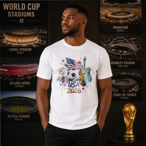 May include: A white short-sleeve t-shirt featuring a graphic print of the American flag, a bald eagle, a soccer ball, a gold trophy, and the Statue of Liberty, with the text USA 2026 printed in a bold, stylized font below the central design.