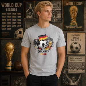 May include: A light gray short-sleeved t-shirt featuring a graphic print of a soccer ball, the German flag, a black eagle, the Brandenburg Gate, and a gold trophy. The text "GERMANY 2026" is printed in black and yellow below the central graphic.