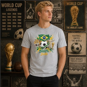 May include: A heather gray t-shirt featuring a graphic print of a soccer ball, a jaguar, the Brazilian flag, the Christ the Redeemer statue, and the World Cup trophy, with the text "BRAZIL 2026" printed in yellow and green below the central design.
