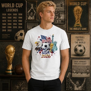 K&ouml;nnte beinhalten: Ein hellgraues T-Shirt mit Rundhalsausschnitt und einem Grafikdruck, der einen Fu&szlig;ball, die amerikanische Flagge, einen Wei&szlig;kopfseeadler, die Freiheitsstatue und einen goldenen Pokal zeigt, erg&auml;nzt durch Farbspritzer und den Text USA 2026.