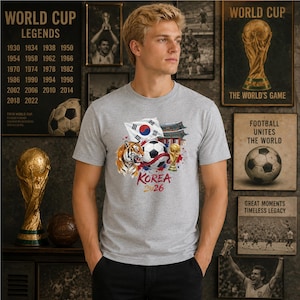 May include: A heather gray t-shirt featuring a graphic print of a soccer ball, a tiger, the South Korean flag, a traditional building, and a gold trophy. The text KOREA 2026 is printed in red and gold below the central soccer ball illustration.