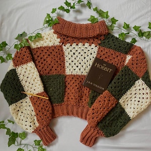 May include: A crocheted sweater with a turtleneck collar in a patchwork design of brown, cream, and green squares. The sweater is laying on a white surface with green ivy vines.