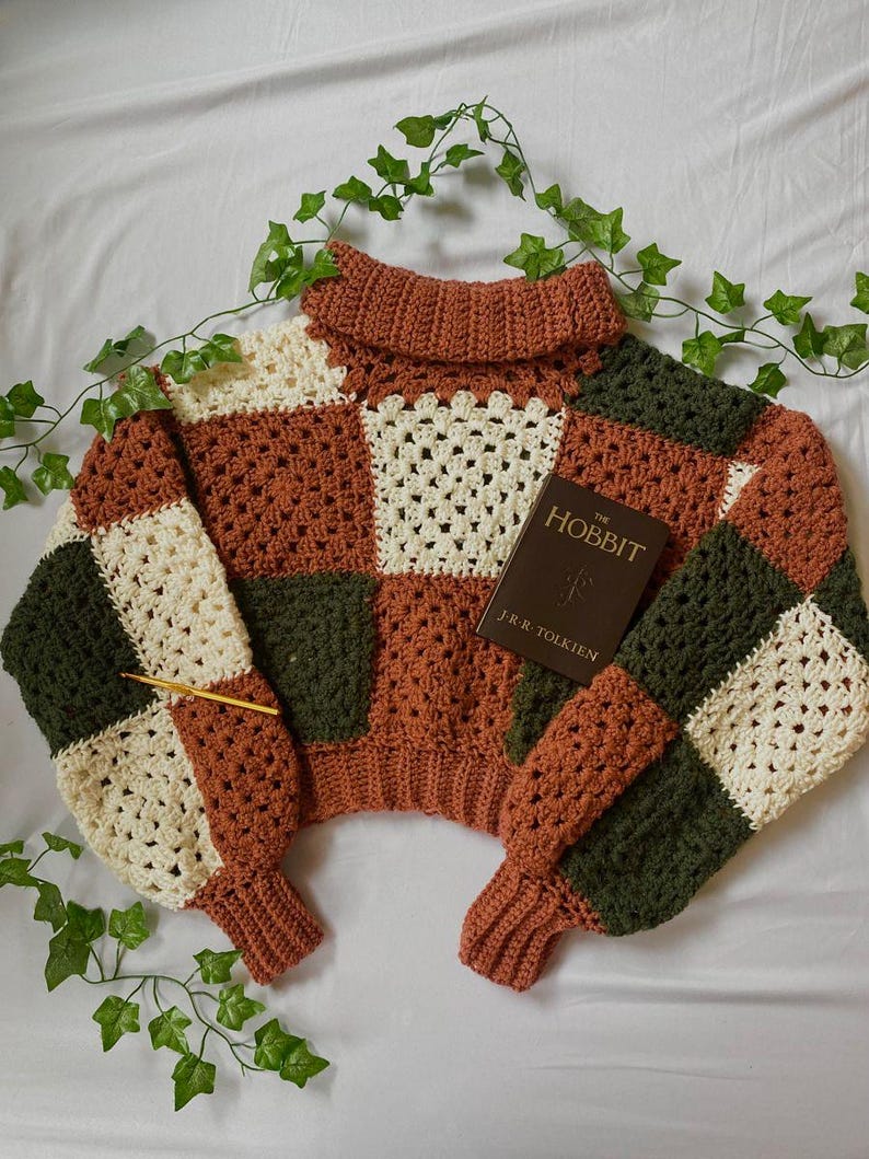 May include: A crocheted sweater with a turtleneck collar in a patchwork design of brown, cream, and green squares. The sweater is laying on a white surface with green ivy vines.