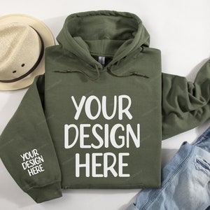 Pode incluir: Uma camisola de capuz verde com o texto "YOUR DESIGN HERE" impresso em branco na frente. A camisola est&aacute; colocada sobre uma superf&iacute;cie branca com um chap&eacute;u de palha e cal&ccedil;as de ganga azuis.
