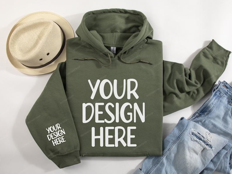 Pode incluir: Uma camisola de capuz verde com o texto "YOUR DESIGN HERE" impresso em branco na frente. A camisola est&aacute; colocada sobre uma superf&iacute;cie branca com um chap&eacute;u de palha e cal&ccedil;as de ganga azuis.