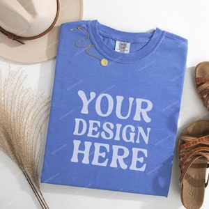 May include: A blue t-shirt with the text "YOUR DESIGN HERE" printed on the front. The shirt is laid flat on a white surface with a straw-like plant, a hat, and sandals.