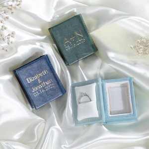 May include: Three small, book-shaped ring boxes. One is blue with the text "Elizabeth & Jonathan 03.16.2025", one is green with the text "A+N 03.16.2025", and one is light blue with a ring inside.