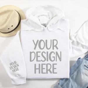 Pode incluir: Camisola de moletom branca com capuz com o texto "YOUR DESIGN HERE" na frente e "YOUR DESIGN HERE" na manga. A camisola est&aacute; colocada sobre uma superf&iacute;cie branca com um chap&eacute;u de palha, uma cal&ccedil;a jeans azul e uma camiseta branca.