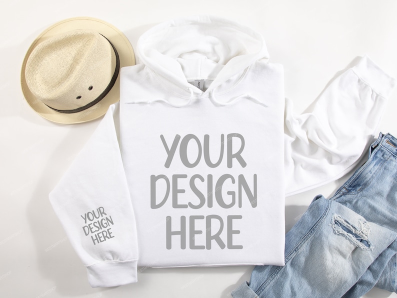 Pode incluir: Camisola de moletom branca com capuz com o texto "YOUR DESIGN HERE" na frente e "YOUR DESIGN HERE" na manga. A camisola est&aacute; colocada sobre uma superf&iacute;cie branca com um chap&eacute;u de palha, uma cal&ccedil;a jeans azul e uma camiseta branca.