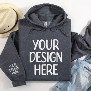 Pode incluir: Uma camisola de capuz cinzenta escura com o texto "YOUR DESIGN HERE" impresso em branco na frente. A camisola est&aacute; colocada sobre uma superf&iacute;cie branca com um chap&eacute;u de palha e cal&ccedil;as de ganga azuis.