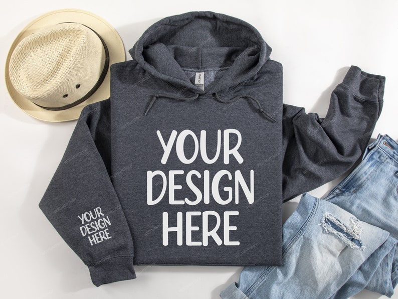 Pode incluir: Uma camisola de capuz cinzenta escura com o texto "YOUR DESIGN HERE" impresso em branco na frente. A camisola est&aacute; colocada sobre uma superf&iacute;cie branca com um chap&eacute;u de palha e cal&ccedil;as de ganga azuis.
