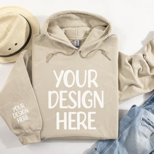 Pode incluir: Um moletom bege com cord&atilde;o branco e o texto "YOUR DESIGN HERE" estampado na frente. O moletom est&aacute; colocado sobre uma superf&iacute;cie branca com um chap&eacute;u de palha e uma cal&ccedil;a jeans azul.