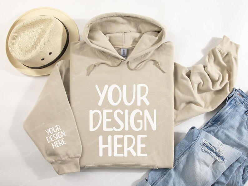 Pode incluir: Um moletom bege com cord&atilde;o branco e o texto "YOUR DESIGN HERE" estampado na frente. O moletom est&aacute; colocado sobre uma superf&iacute;cie branca com um chap&eacute;u de palha e uma cal&ccedil;a jeans azul.