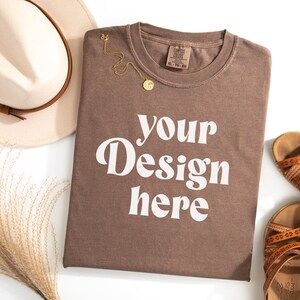 May include: A brown t-shirt with the text "your design here" in white. The t-shirt is folded and laid flat on a white surface. There is a hat, a pair of sandals, and a sprig of dried grass in the background.