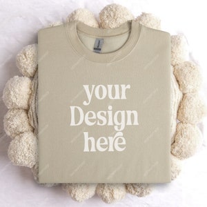 May include: A beige sweatshirt with the text "your design here" printed on the front. The sweatshirt is laid flat on a white surface with a cream-colored pom-pom rug.