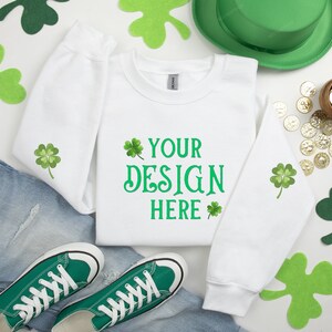 Puede incluir: Sudadera blanca con diseños de trébol verde y el texto "YOUR DESIGN HERE". La sudadera está colocada sobre una superficie blanca con tréboles verdes, un sombrero verde y monedas de oro.