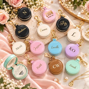 May include: A collection of round, zippered jewelry cases with gold-tone metal clasps, available in black, white, pink, blue, purple, and brown. Each case features personalized gold lettering, including names like Madison, Emma, and Zion National Park.