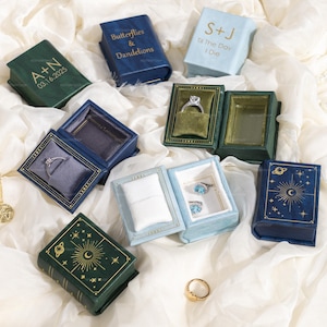 May include: A collection of small, book-shaped ring boxes in various colors, including green, blue, and light blue. Some boxes are open, displaying rings. The boxes have gold-colored text and celestial designs.