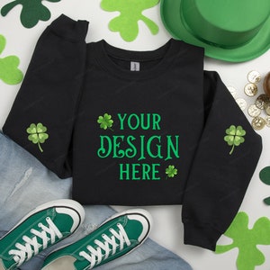 May include: Black sweatshirt with green shamrock designs on the sleeves and the text "YOUR DESIGN HERE" on the front. The sweatshirt is laid out on a white surface with green shamrocks, a green hat, and gold coins.