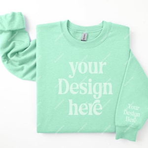May include: A light green sweatshirt with the text "your design here" printed on the front. The sweatshirt is folded and laid flat on a white surface.