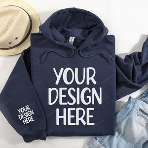 Pode incluir: Um moletom azul marinho com cord&atilde;o branco e o texto "YOUR DESIGN HERE" estampado na frente. O moletom est&aacute; colocado sobre uma superf&iacute;cie branca com um chap&eacute;u de palha e uma cal&ccedil;a jeans azul.