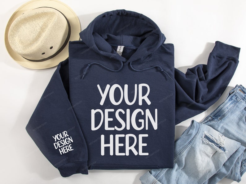 Pode incluir: Um moletom azul marinho com cord&atilde;o branco e o texto "YOUR DESIGN HERE" estampado na frente. O moletom est&aacute; colocado sobre uma superf&iacute;cie branca com um chap&eacute;u de palha e uma cal&ccedil;a jeans azul.