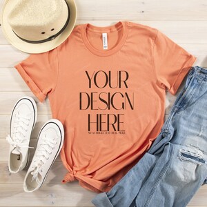 May include: A peach coloured t-shirt with the text "YOUR DESIGN HERE NO AL TRICKS, JUST REAL PICKS" printed on the front. The t-shirt is laid out on a wooden surface with a pair of white trainers, a straw hat, and a pair of blue jeans.