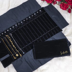 May include: A black velvet jewelry roll with multiple compartments, holding gold and silver necklaces. A smaller matching pouch is next to the roll, with the name "Isabella" in gold script. The roll is open, displaying the necklaces.