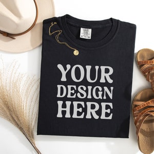 May include: Black t-shirt with the text "YOUR DESIGN HERE" in white. The t-shirt is folded and laid flat on a white surface. There is a straw hat, a necklace, and sandals in the background.