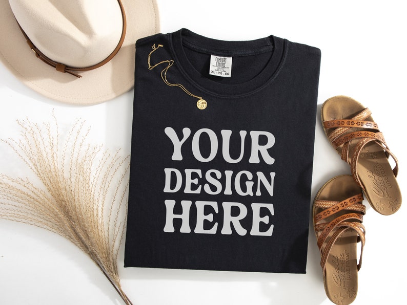 May include: Black t-shirt with the text "YOUR DESIGN HERE" in white. The t-shirt is folded and laid flat on a white surface. There is a straw hat, a necklace, and sandals in the background.