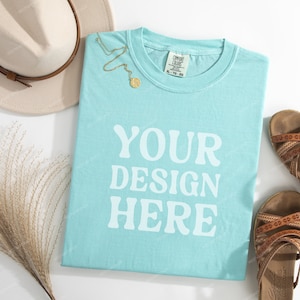 May include: A light blue t-shirt with the text "YOUR DESIGN HERE" in white. The shirt is laying flat on a white surface with a straw hat, a gold necklace, and two pairs of brown sandals.