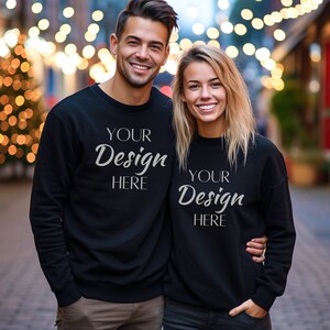 Puede incluir: Dos sudaderas negras con el texto "Your Design Here" en la parte delantera. Las sudaderas las lleva puestas una pareja que está de pie frente a un fondo borroso de luces navideñas.